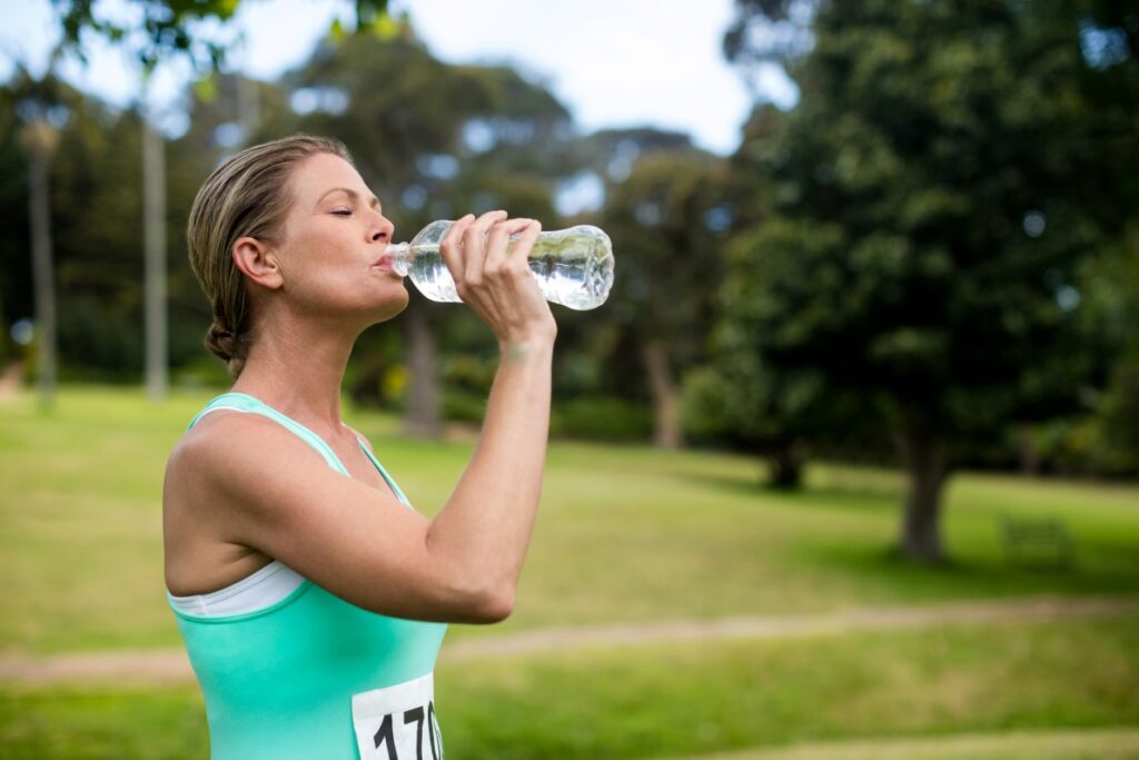 Woman in tank top with a marathon number on the front drinks from a bottle of water during her race. She will need to find the best way to rehydrate in Nashville after the race to support her recovery.